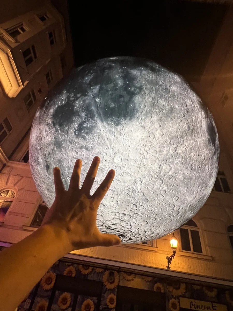 Kivilágított, óriási holdinstalláció egy szűk utca házai fölött, alulról fényképezve, egy kinyújtott kézzel az előtérben.